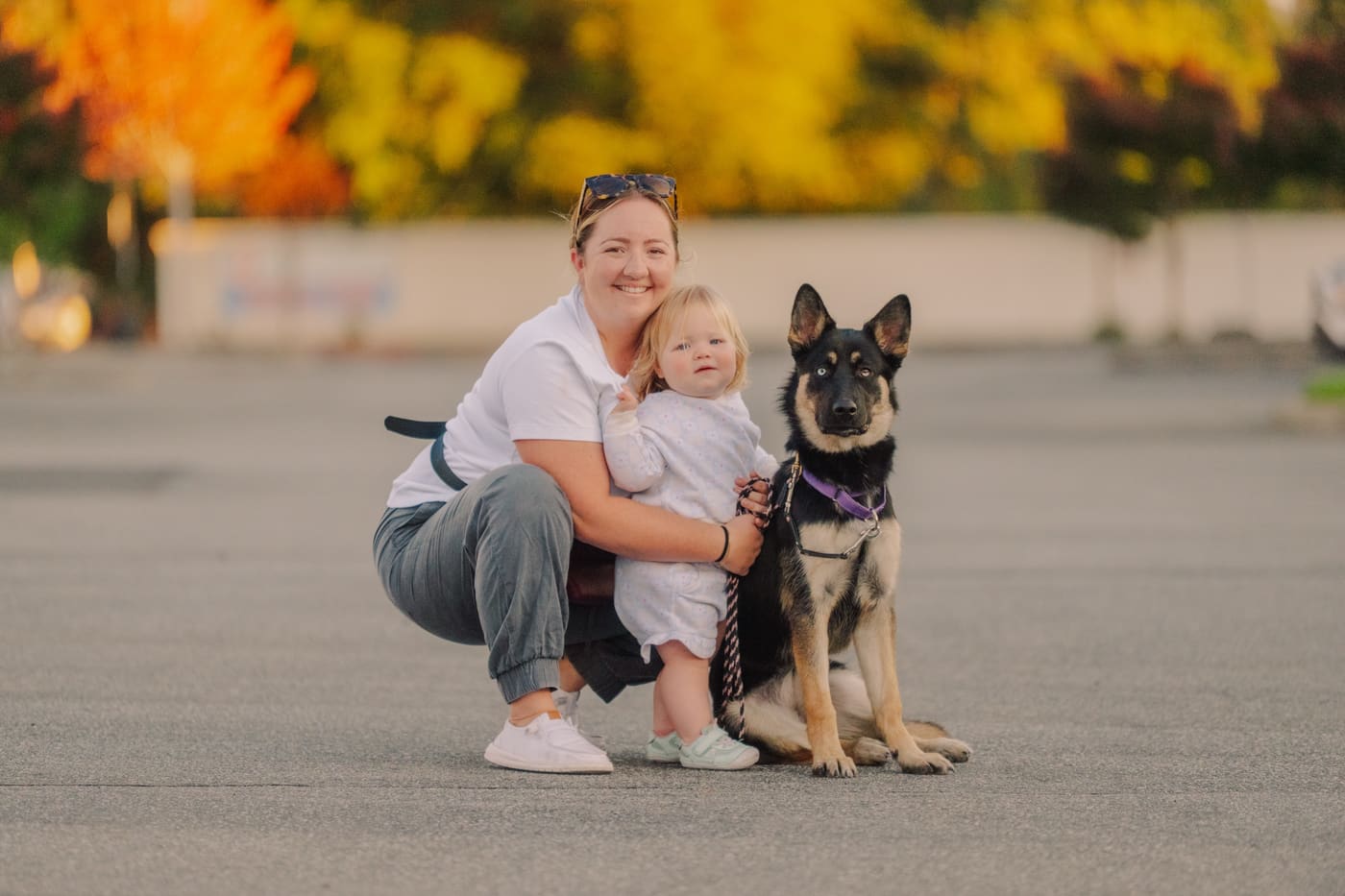 A mother and toddler crouched beside their well-trained German Shepherd outdoors